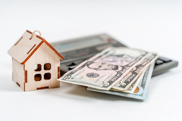 Un modèle en bois de la maison se dresse avec une calculatrice et des billets d'un dollar sur fond blanc