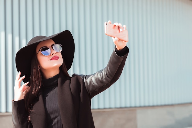 Le modèle brune élégant porte un chapeau et des lunettes de soleil à la mode, prenant le selfie sur le téléphone portable