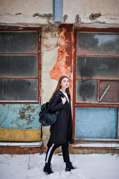 Modèle brune à la mode à longues jambes dans une longue cape noire posée en plein air le jour d'hiver contre un vieux mur de grunge avec des fenêtres cassées.