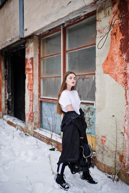 Modèle brune à la mode à longues jambes dans une longue cape noire posée en plein air le jour d'hiver contre un vieux mur de grunge avec des fenêtres cassées.