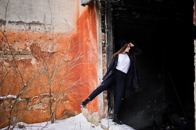 Modèle brune à la mode à longues jambes dans une longue cape noire posée en plein air le jour d'hiver contre le vieux mur de grunge.