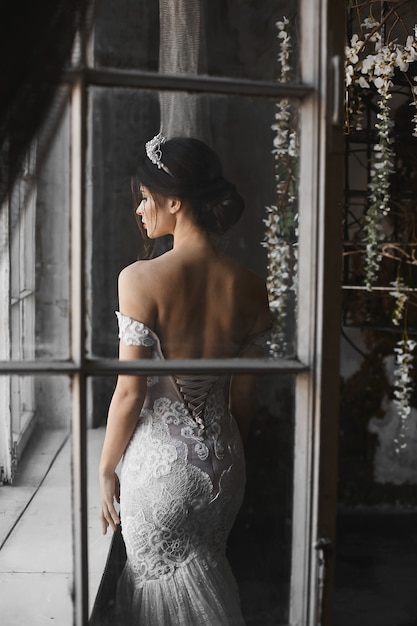 Modèle brune en robe blanche en dentelle avec épaules nues avec diadème sur la tête posant près de la fenêtre