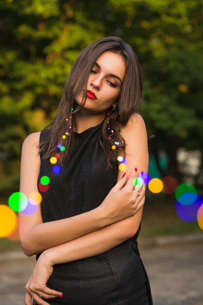 Modèle brune sensuelle avec un maquillage lumineux tenant une guirlande dans le parc de la ville