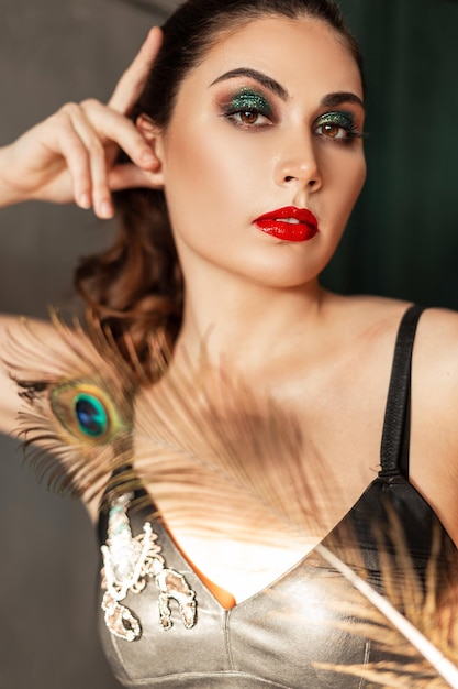 Modèle élégant de belle jeune femme avec le maquillage lumineux, les lèvres rouges et les paupières vertes dans des vêtements à la mode avec une plume de paon regarde la caméra dans le studio