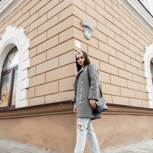 Modèle élégant jolie jeune femme en bandana à la mode en manteau élégant gris avec sac à main noir posant près d'un bâtiment vintage à l'extérieur. Belle fille dans des vêtements de printemps décontractés pour les jeunes à la mode aime marcher.