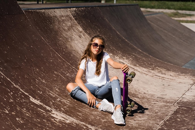 Modèle enfant dans des verres assis et posant avec penny board sur l'aire de skate Enfant fille active avec profiter d'un style de vie extrême sur la rampe de sport