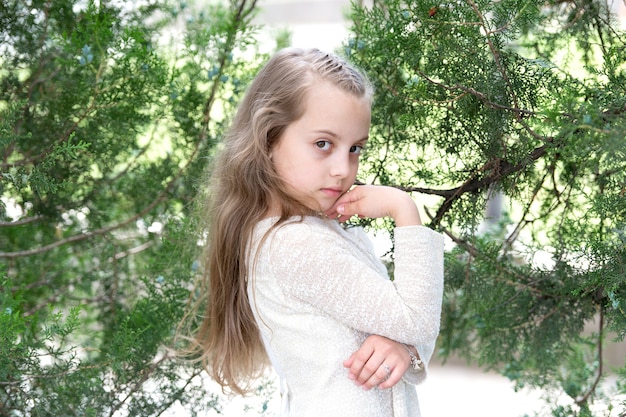 Modèle d'enfant avec une peau de visage fraîche dans la nature estivale. Fille en robe de mode pose à l'arbre vert. Petit enfant aux longs cheveux blonds en plein air. Mode beauté et coiffure. Soin et fraîcheur.