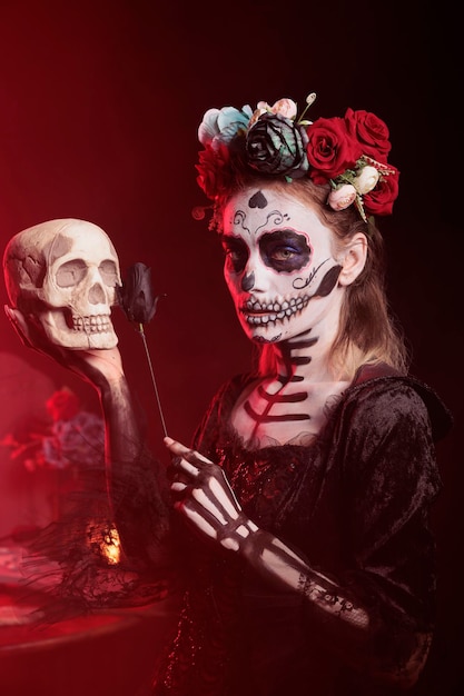 Modèle fantasmagorique tenant le crâne et la rose noire en studio, agissant comme l'horreur la cavalera catrina en costume d'halloween. Dame de la mort célébrant la tradition des fêtes mexicaines, le jour des morts.