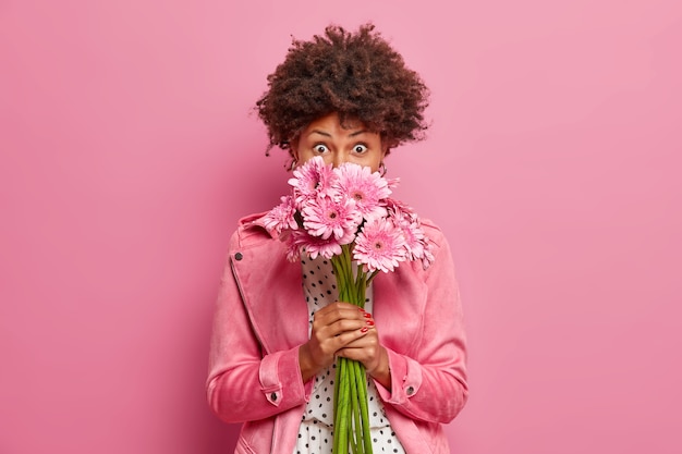 Un modèle féminin afro-américain aux cheveux bouclés doux sent un bouquet de gerberas roses détient de belles fleurs reçues pour un anniversaire ou une occasion spéciale vêtue d'une veste élégante pose sur un mur rose