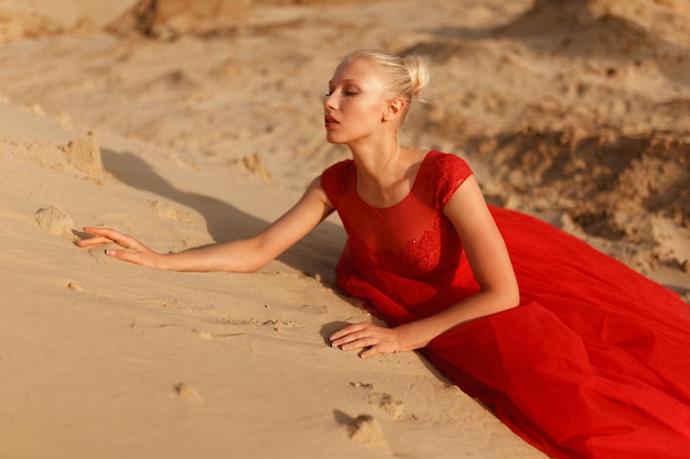 Le modèle féminin blond sensuel dans une belle robe rouge élégante se trouve sur le sable.