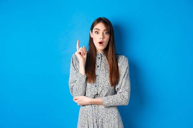 Modèle féminin excité en robe levant le doigt, présentant une idée et un regard étonné devant la caméra, a trouvé une solution ou a planifié, compris quelque chose, debout sur le bleu.