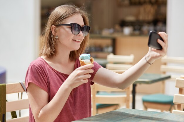 Modèle féminin gai avec un sourire amical mange de la crème glacée et fait un portrait de selfie sur téléphone portable