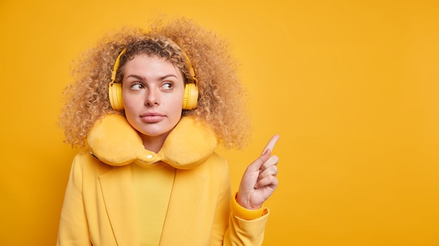 Photo modèle féminin millénaire aux cheveux bouclés sérieux donne une recommandation indique à l'espace de copie vierge porte des écouteurs stéréo sur les oreilles oreiller de voyage autour du cou isolé sur mur jaune