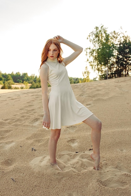 Modèle féminin mince en robe d'été blanche avec de longs cheveux roux debout sur une colline de sable en été