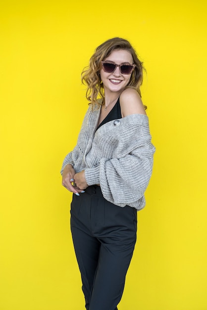 Modèle féminin positif posant en pull tricoté gris à lunettes de soleil près du mur jaune, extérieur