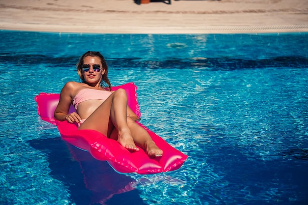 Modèle féminin sexy se reposant et prenant un bain de soleil sur un matelas dans la piscine Femme en maillot de bain bikini rose flottant sur un matelas rose gonflable spf et crème solaire