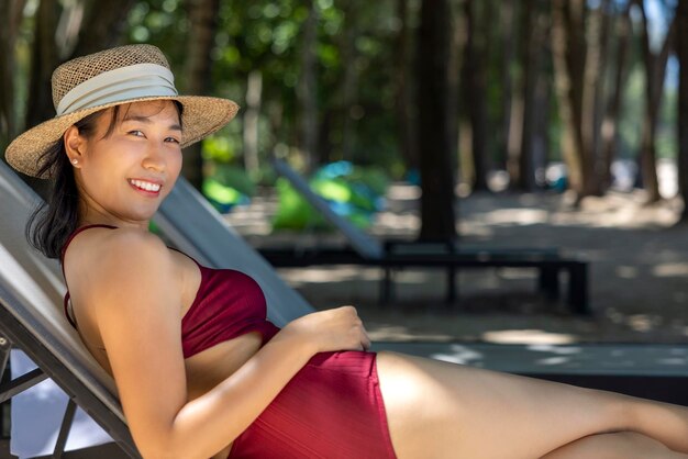 Modèle femme asiatique portant un maillot de bain rouge et un chapeau de paille allongé sur la chaise longue au soleil au bord de la mer
