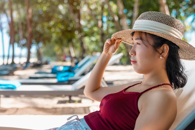 Modèle femme asiatique portant un maillot de bain rouge et un chapeau de paille est assis au soleil au bord de la mer