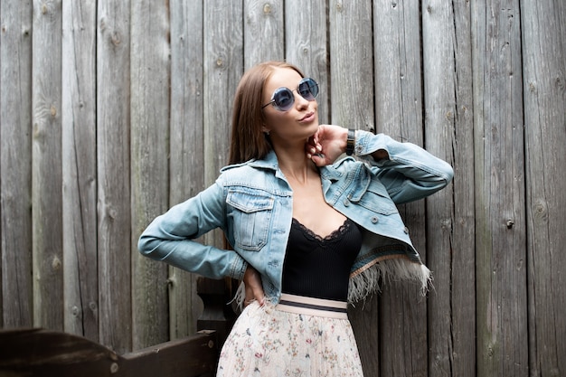Modèle de femme assez glamour avec des lunettes de soleil dans des vêtements en denim à la mode avec de la lingerie en dentelle noire avec une jupe se tient près d'un mur en bois à la campagne