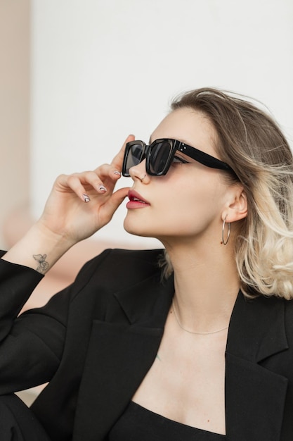 Modèle de femme élégante jeune entreprise à la mode avec des lunettes de soleil modernes à la mode en tenue noire avec haut et blazer est assis dans la rue Vêtements féminins urbains