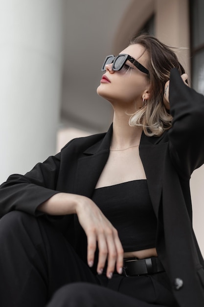 Modèle de femme de mode d'affaires magnifiquement élégant avec des lunettes de soleil élégantes modernes dans des vêtements à la mode noirs avec un haut blazer noir et un pantalon se trouve dans la ville