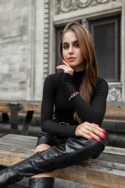 Modèle de femme à la mode cool dans une tenue d'été noire à la mode avec un short de golf à manches longues et des chaussures hautes est assis sur un banc en bois près d'un bâtiment vintage