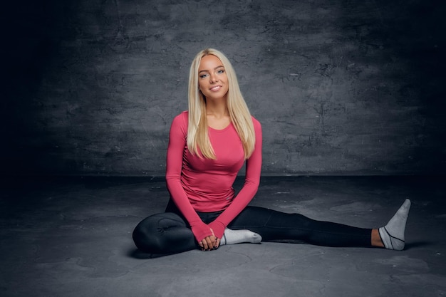 Un modèle de fitness blond positif et flexible est assis sur un sol dans un studio sur fond gris.