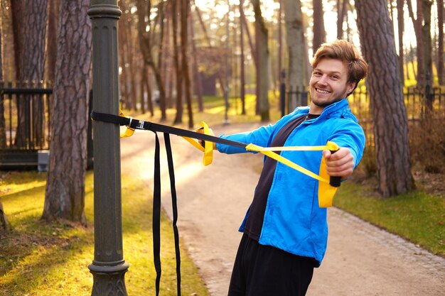 Modèle de fitness homme dans une veste de sport bleue faisant des séances d'entraînement avec des bandes trx de fitness dans un parc.