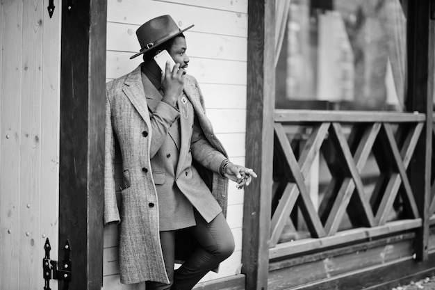 Modèle d'homme afro-américain élégant en manteau gris veste cravate et chapeau rouge posé contre un café en bois et parlant au téléphone mobile Photo noir et blanc