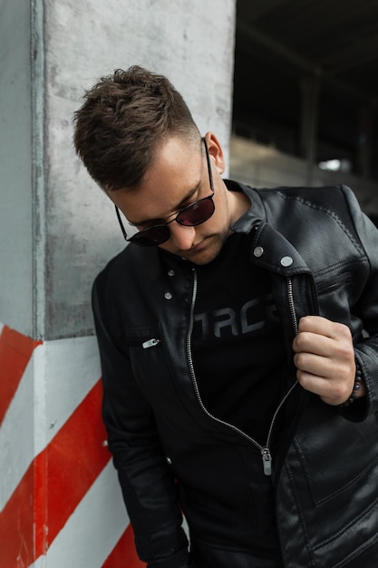 Modèle d'homme de rue élégant avec des cheveux avec des lunettes de soleil noires portant des vêtements noirs avec une veste en cuir noire dans la ville Vêtements de style masculin urbain