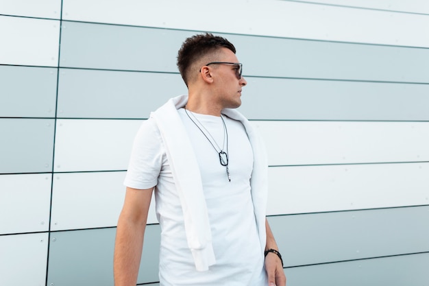 Modèle d'homme urbain à la mode dans des lunettes de soleil noires élégantes dans un t-shirt à la mode blanc se dresse près d'un mur moderne de la ville.