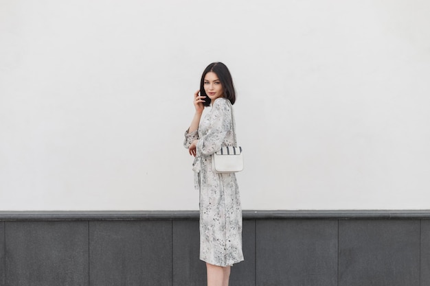 Modèle de jeune femme brune à la mode avec une coiffure short en robe de fleurs de printemps vintage élégante avec sac à main de mode marche et se tient dans la rue près d'un mur blanc