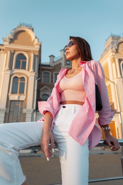Modèle de jeune femme à la mode à la mode avec des lunettes élégantes dans une chemise rose à la mode, un débardeur, un jean blanc et un sac à main se trouve dans la ville Look de style féminin urbain d'été