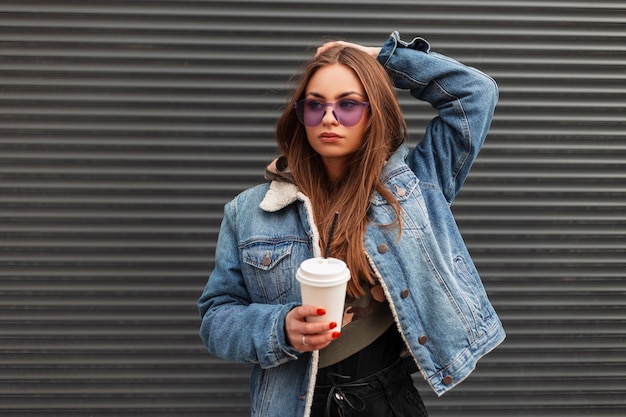 Modèle de jeune femme urbaine glamour dans des lunettes violettes à la mode dans une veste en jean élégante avec une tasse de boisson chaude posant près d'un mur métallique à l'extérieur. Une jolie fille hipster avec du café profite de la journée de printemps.Style