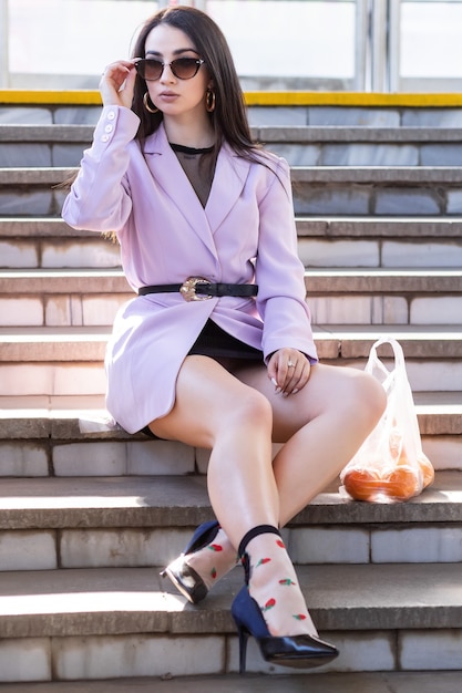 Le modèle de jolie femme de mode avec des oranges de fruits est assis sur les marches dans une veste violette, des chaussures noires.