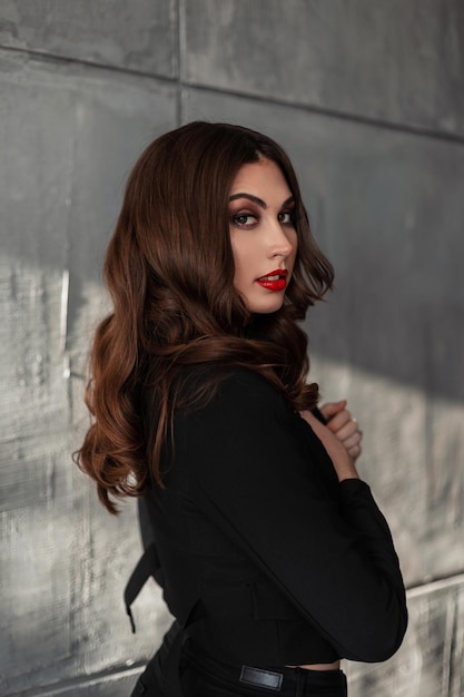 Modèle de jolie jeune femme avec du maquillage et des cheveux bouclés dans des vêtements de costume noir à la mode près d'un mur de béton gris à l'intérieur
