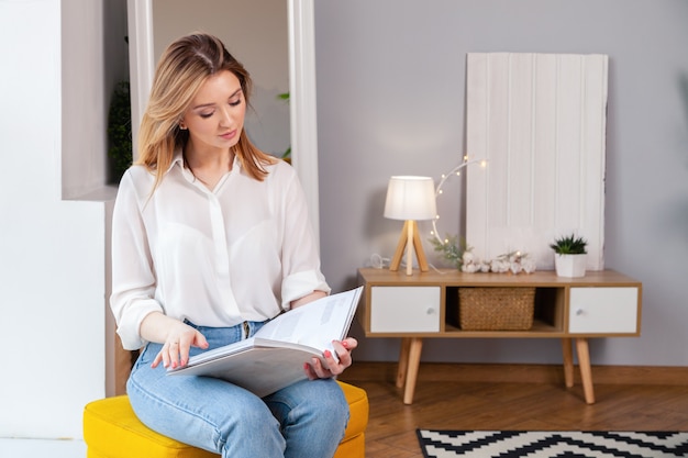 Modèle jolie jolie jolie fille caucasien en chemise blanche, jeans bleu assis sur une chaise et un magazine de lecture.