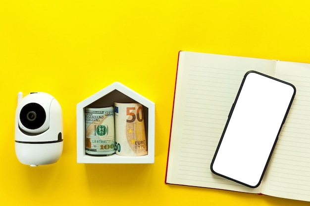 Modèle d'une maison blanche en bois avec des factures papier, un téléphone et une caméra de sécurité sur fond jaune