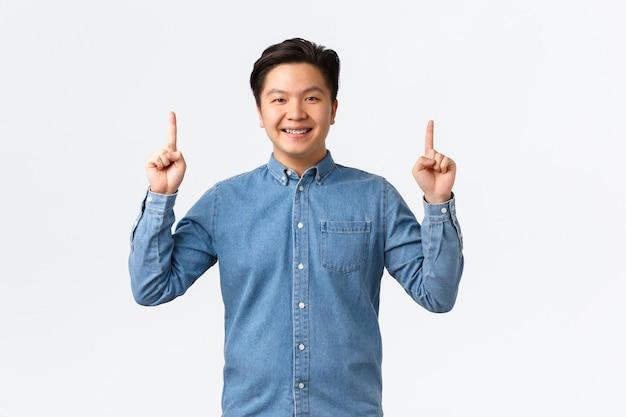 Modèle masculin asiatique souriant, fier et heureux en chemise bleue faisant une annonce, pointant les doigts vers le haut sur la bannière, recommande d'acheter le produit, cliquez sur le lien, concept de commerce électronique et de publicité