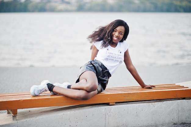 Modèle mince afro-américain à la peau foncée posé dans un short en cuir noir et un t-shirt blanc Elle luying sur un banc