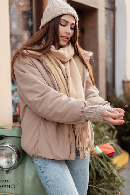 Modèle à la mode de jolie jeune femme en vêtements d'extérieur élégants avec un bonnet tricoté, un pull beige, une veste et une écharpe se tient près d'un scooter rétro dans la ville. Vacances et voyages