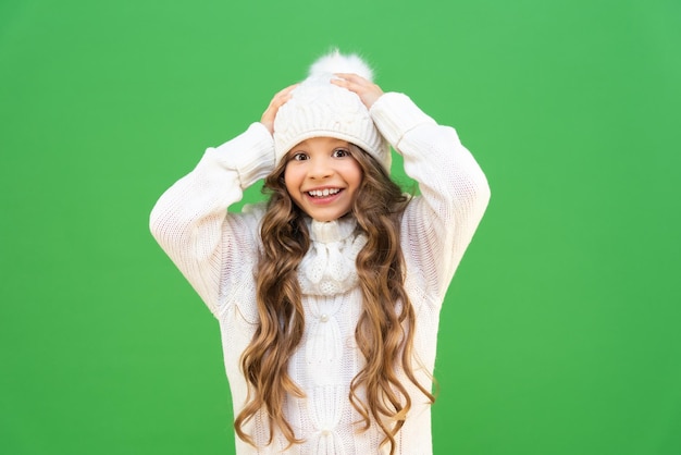 Un modèle de petite fille tient un chapeau d'hiver et sourit. Bébé aux cheveux bouclés et un beau sourire dans un chapeau chaud et un pull tricoté sur un fond vert isolé.