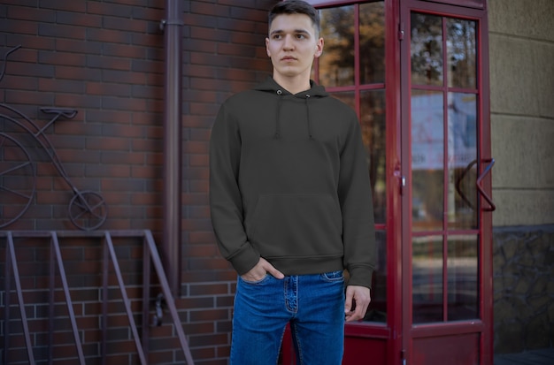 Modèle De Sweat à Capuche Noir Sur Un Jeune Homme, Vue De Face, Présentation De Vêtements Dans La Rue. Maquette De Vêtements Pour Hommes Vides Pour La Publicité Dans La Boutique En Ligne. Capuche Avec Manches Pour Votre Conception