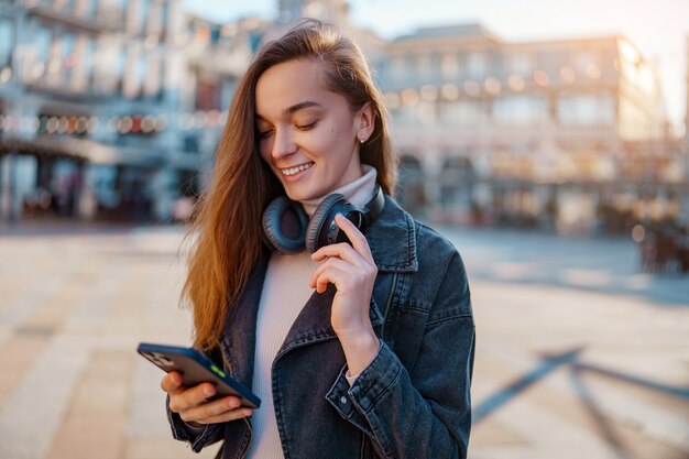 Modern smiling happy casual jeune femme à l'aide d'écouteurs sans fil et smartphone à l'extérieur