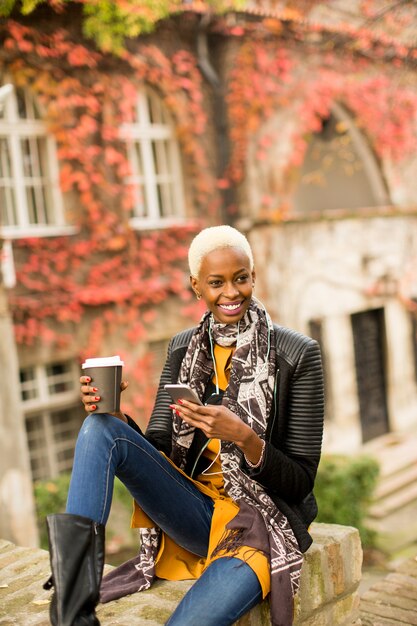 Moderne jeune femme afro-américaine posant avec café pour aller et téléphone mobile en plein air