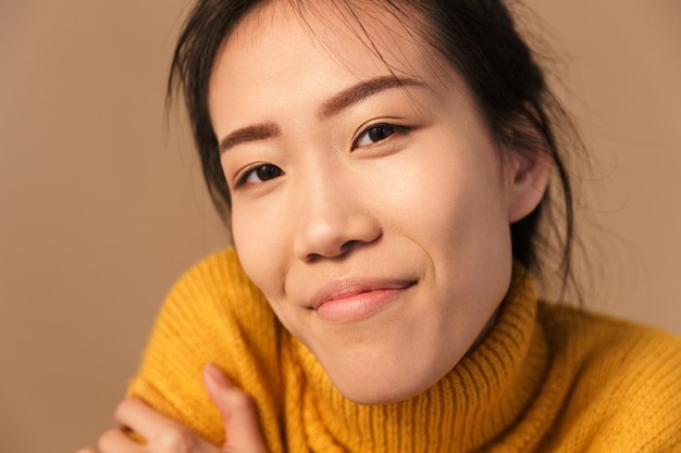 modeste femme asiatique portant un pull souriant tout en posant devant en studio isolé sur un mur beige