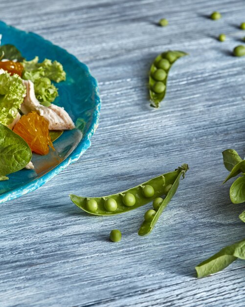 Photo la moitié d'une assiette en céramique bleue avec salade maison à partir d'ingrédients naturels - légumes verts frais, viande de poulet, agrumes, gousses de pois sur une table en bois gris.