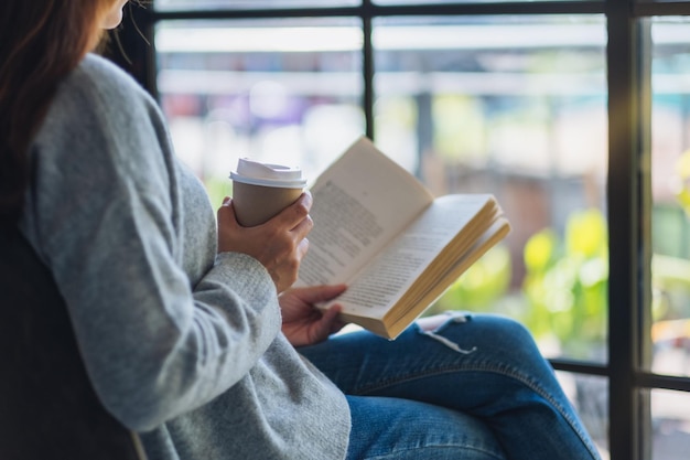 La moitié d'une femme qui prend du café en lisant un livre sur une chaise contre la fenêtre