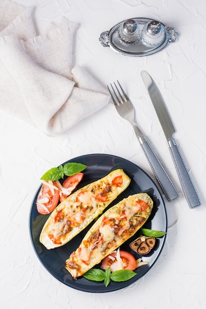 Moitiés de courgettes cuites au four prêtes à manger remplies de fromage et de feuilles de tomates et de basilic et de couverts sur une plaque noire sur un tableau blanc. Menu de légumes, alimentation saine. Vue de dessus et verticale