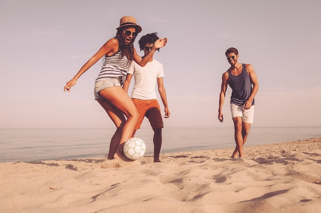 Moment amusant avec des amis. Trois jeunes joyeux jouant avec un ballon de football sur la plage